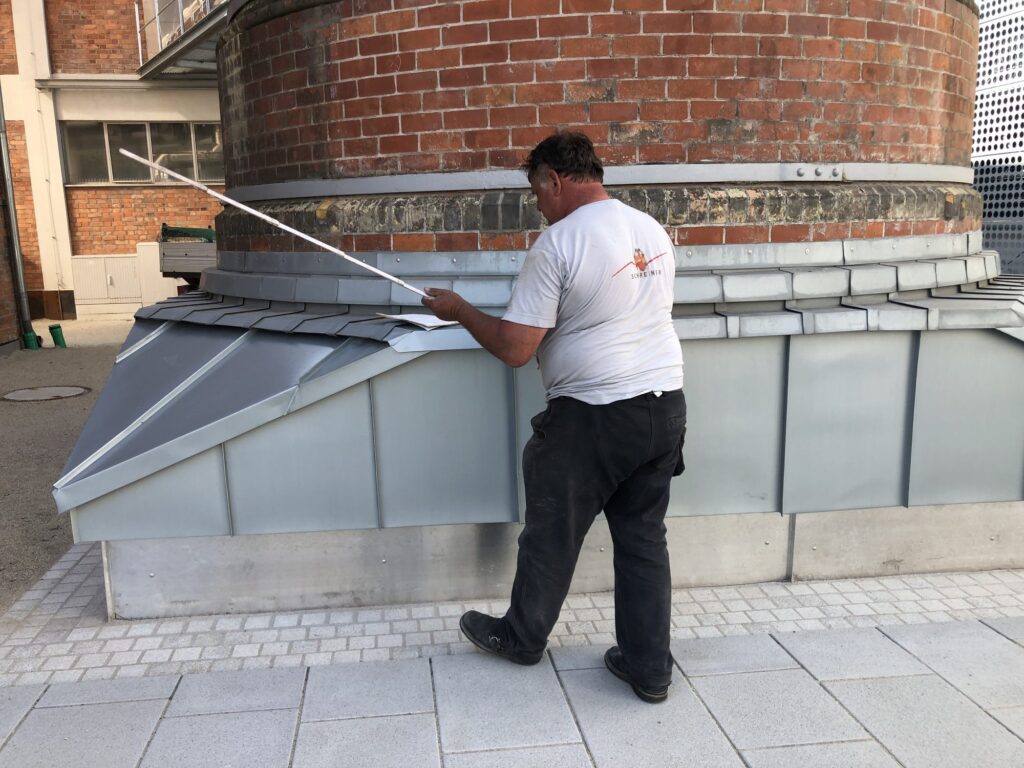 Mitarbeiter misst die Blechverkleidung am Sockel eines runden Backsteinbaus